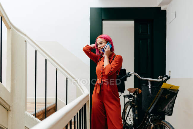 Young stylish female in red suit with backpack talking on smartphone while standing with bicycle on staircase — Stock Photo