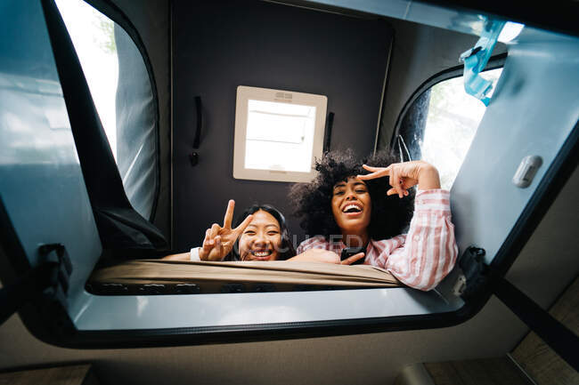 From below of cheerful young diverse female friends showing peace sign and looking at camera through window inside camper van while having fun together during summer vacation — Stock Photo