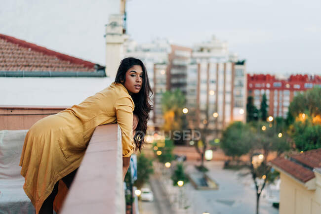 Junge hispanische Frau in lässiger Kleidung lehnt am Geländer und blickt in die Kamera, während sie es sich abends auf dem Balkon in der Stadt gemütlich macht — Stockfoto