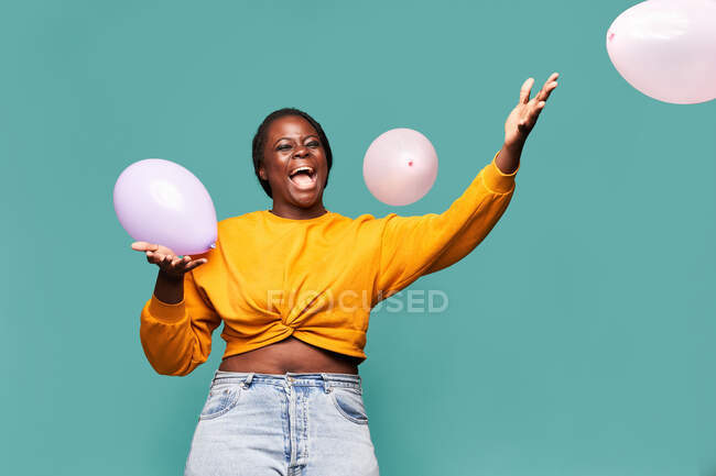 Excited African American female in jeans and yellow top standing near falling balloons against blue background in studio — Stock Photo