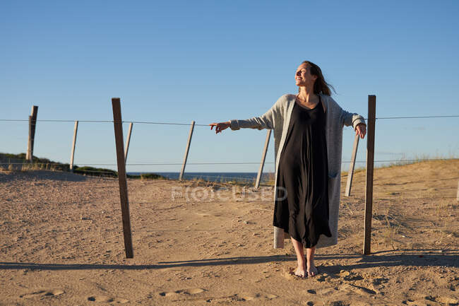 Задоволена жінка в одязі стоїть біля паркану на піщаному пляжі і насолоджується сонячним днем влітку — стокове фото