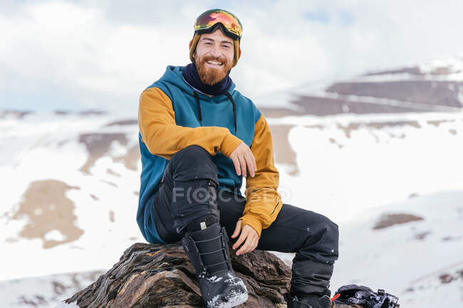 Atleta masculino barbudo sonriente en ropa deportiva mirando a la cámara mientras está sentado en el monte en temporada de invierno en España - foto de stock