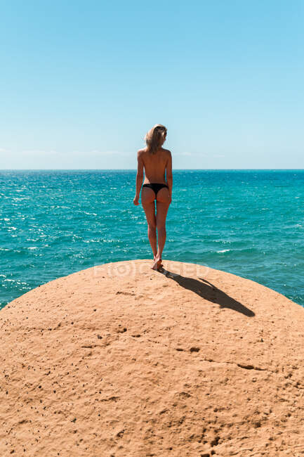 Ganzkörperrückansicht einer unkenntlich gewordenen Blondine in Hose, die an der steinigen Küste vor dem ruhigen türkisfarbenen Meer steht und den sonnigen Tag im Sommerurlaub genießt — Stockfoto