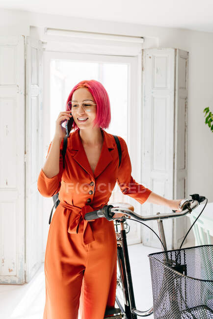 Jovem designer feminina criativa em roupas da moda e óculos falando no smartphone enquanto está de pé com bicicleta no apartamento leve moderno — Fotografia de Stock