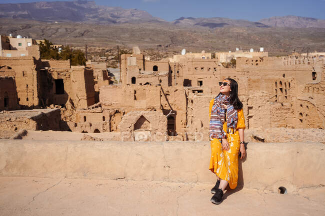 Heureuse jeune touriste féminine en tenue colorée souriant et regardant la caméra alors qu'elle était assise sur des bâtiments anciens en ruine du village abandonné d'Al Hamra à Oman — Photo de stock