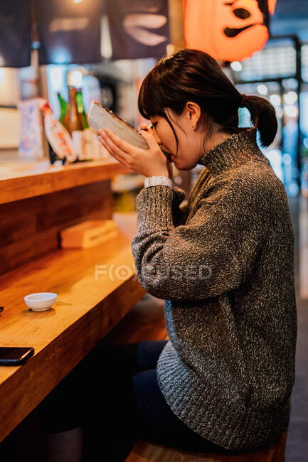 Young Asian woman in sweater eating ramen with spoon at wooden counter in cafe — Stock Photo