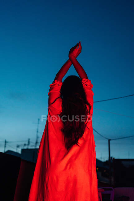 Вид ззаду на невизначену етнічну жінку в нижній білизні, дивлячись на камеру, відпочиваючи на терасі під червоним неоновим світлом вночі з синім темним небом — стокове фото