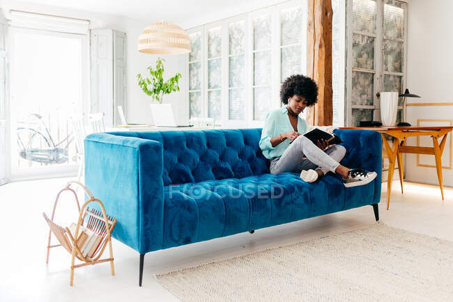Молодая черная женщина в повседневной одежде с наушниками сидит на уютном синем диване дома и читает журнал, наслаждаясь свободным временем дома — стоковое фото
