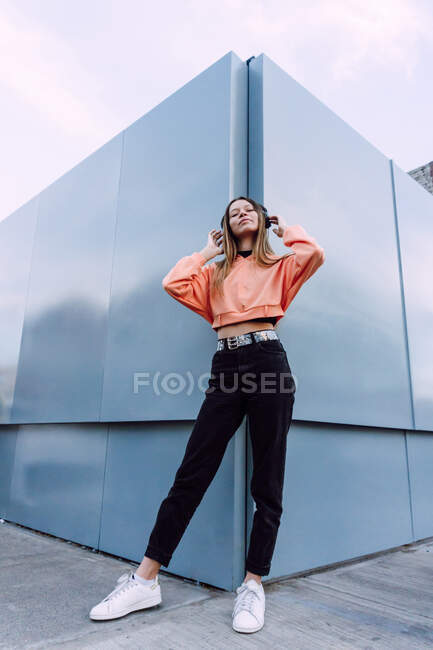 Low angle full body of stylish female standing near modern geometric building and listening to music in wireless headphones — Stock Photo