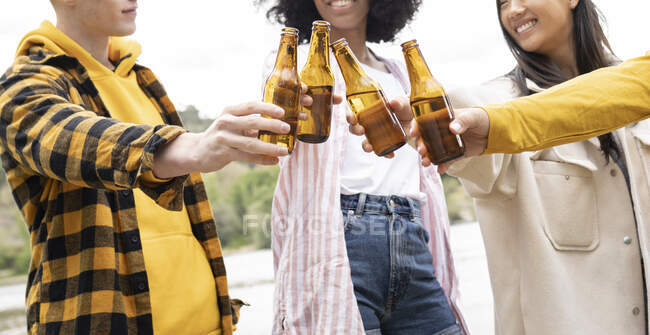 Cropped unrecognizable man and women clinking bottles of beer and proposing toast while resting on shore of river in nature — Stock Photo