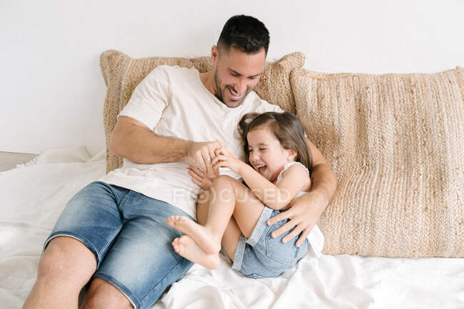 Счастливый отец щекочет смеющуюся маленькую девочку, когда она лежит на кровати и веселится дома — стоковое фото