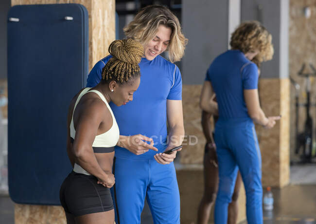 Content young sportsman showing cellphone to black female friend in activewear while interacting in gymnasium — Stock Photo