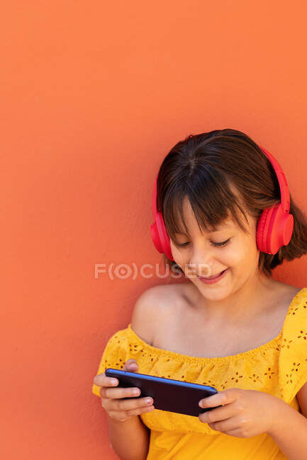Conteúdo criança navegando na internet no celular enquanto ouve música do fone de ouvido sem fio no fundo laranja — Fotografia de Stock