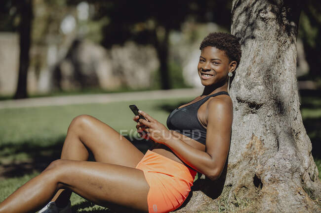 Fröhliche Afroamerikanerin in Sportkleidung hält Smartphone in der Hand, während sie an sonnigen Tagen im Gras sitzt — Stockfoto