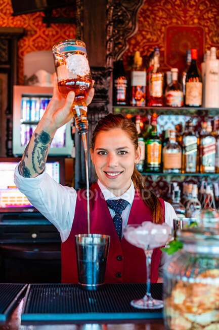 Усмішка самки бармена виливає алкоголь у шейкер, готуючи відсвіжний коктейль за стійкою в барі і дивлячись на камеру. — стокове фото