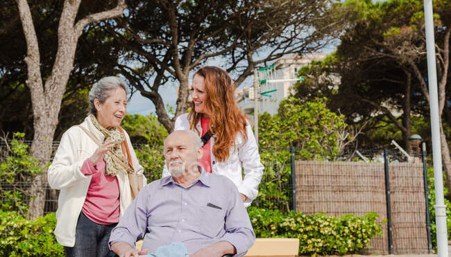 Senior mother and adult daughter talking to each other during stroll with aged father in wheelchair in park on sunny day — Stock Photo