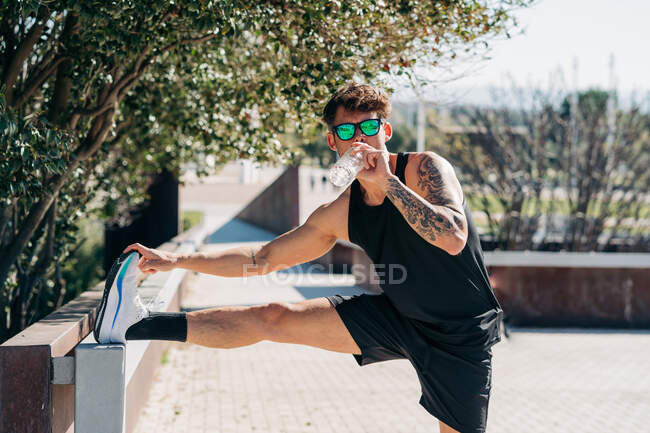 Tattooed male athlete in sportswear working out while drinking aqua from bottle in town on sunny day — Stock Photo
