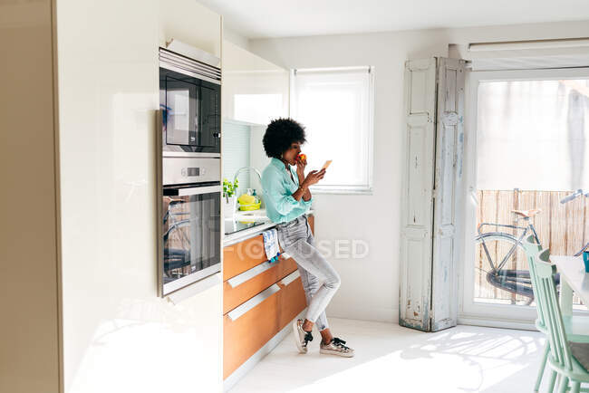 Бічний погляд на молоду афроамериканку в повсякденному одязі з'їдає яблуко і переглядає Інтернет на мобільному телефоні, стоячи вдома на кухні. — стокове фото