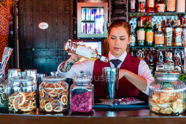 Жінка-бармен, що вливає алкоголь в шейкер, готує освіжаючий коктейль за стійкою в барі і дивиться в сторону — стокове фото