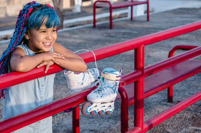 Усміхнена етнічна дитина з різнокольоровими косами, що спираються руками на паркан, дивлячись на тротуар — стокове фото