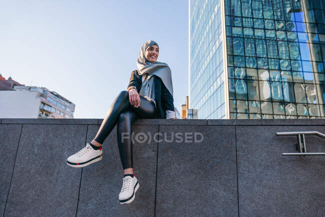 Niedriger Winkel einer lächelnden Muslimin mit Kopftuch und trendiger Kleidung, die auf der Straße am Rand sitzt und wegschaut — Stockfoto