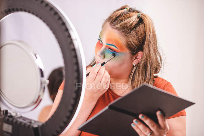Übergewichtige Frau mit Palette, die bunte Pigmente auf das Gesicht aufträgt, während sie im Studio Spiegel in der Nähe von Ringlicht betrachtet — Stockfoto