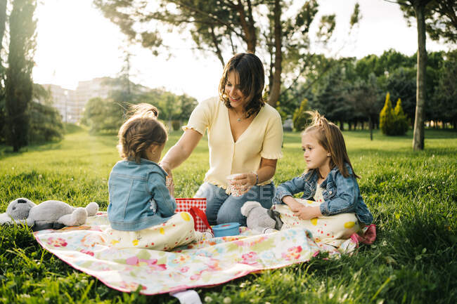 Счастливая молодая женщина с маленькими дочерьми наслаждается пикником на зеленом лугу, проводя вместе летний день в парке — стоковое фото