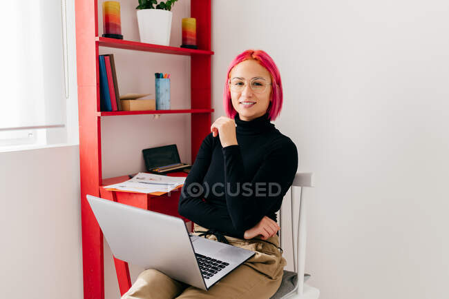 Joven freelancer enfocada en ropa casual sentada en una silla mirando a la cámara usando un portátil mientras trabaja en un proyecto en un apartamento moderno y ligero - foto de stock