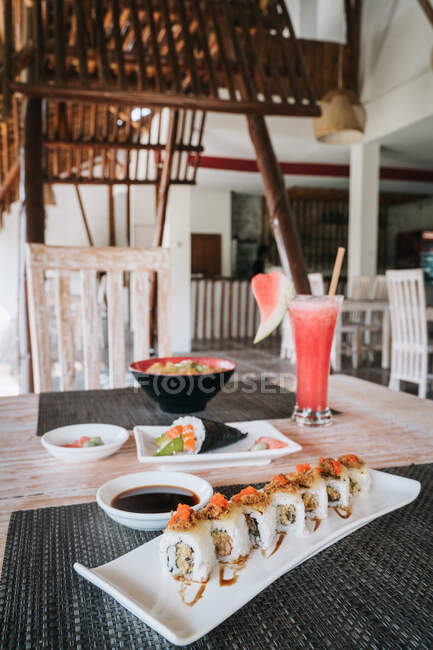 Deliciosa comida asiática de rollos de sushi con salsa de soja contra un tazón y un vaso de bebida en alfombras de mesa - foto de stock