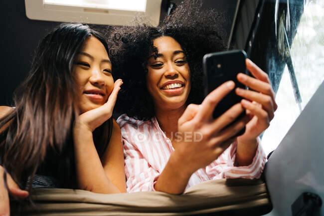 From below of positive young Asian and African American female friends browsing mobile phone while resting together inside camper van during summer trip in nature — Stock Photo