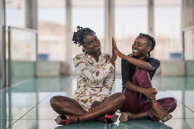 Zufriedene Afroamerikanerinnen, die eng beisammen sitzen und mit nachdenklichem Lächeln in die Kamera schauen, während sie im High-Fiving — Stockfoto