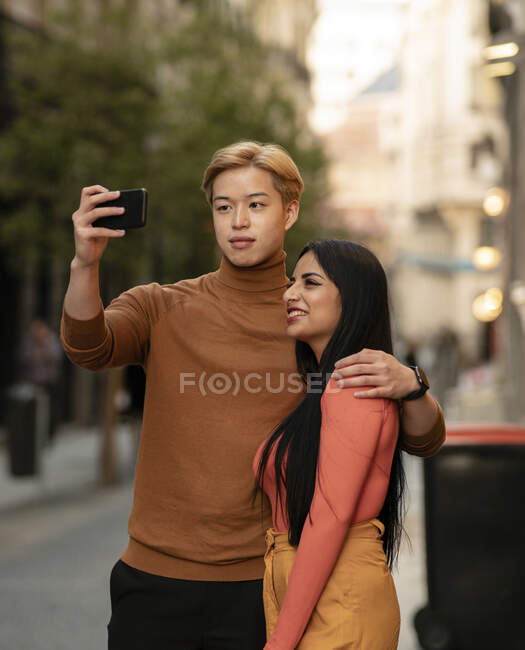 Aimer couple multiethnique étreignant et se prendre en photo sur smartphone tout en se tenant dans la rue — Photo de stock