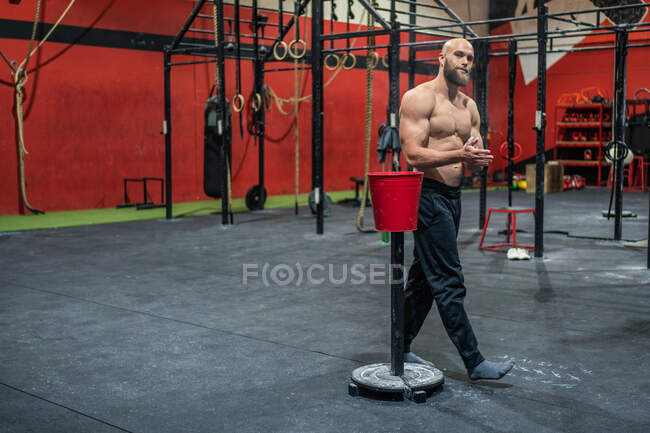 Muscular bearded guy in sportswear looking at camera clapping hands and spreading powder during bodybuilding workout in gym — Stock Photo