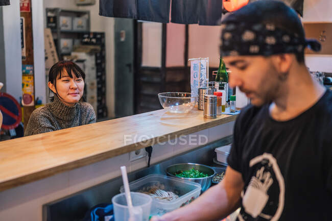 Femme asiatique en tenue décontractée assise au comptoir et parlant avec le travailleur masculin du bar ramen moderne — Photo de stock