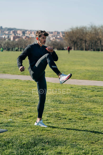 Дорослий спортсмен-чоловік у спортивному одязі, що піднімає ногу і з нетерпінням чекає під час тренувань на газоні на сонячному світлі — стокове фото