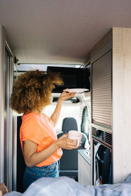 Side view of unrecognizable female traveler with curly hair putting plate into microwave oven while heating food inside modern caravan — Stock Photo