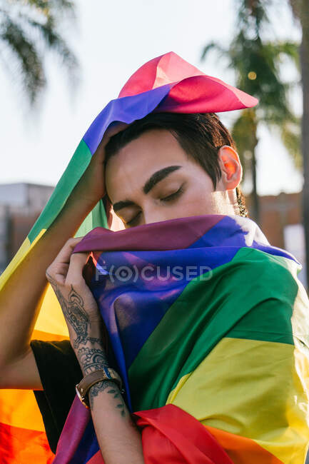 Вид сбоку спокойного гея с закрытыми глазами, завернутым в красочный ЛГБТ-флаг на городской улице — стоковое фото