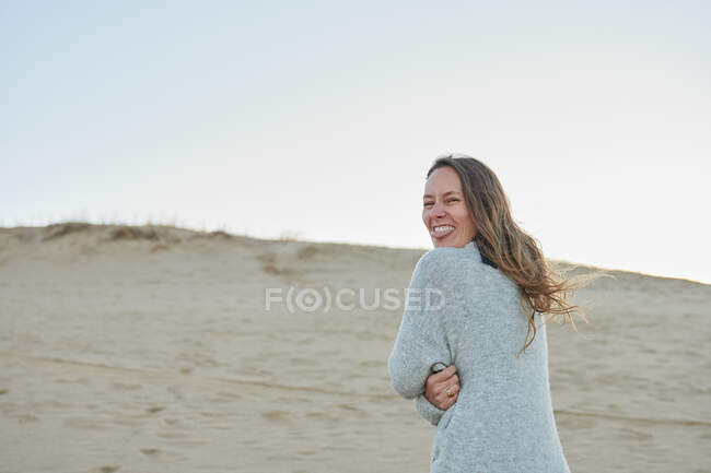 Zufriedene Frau in warmer Kleidung, die in die Kamera blickt, die Zunge am Strand am Meer ausstreckt und den Sommerabend genießt, während sie in die Kamera schaut — Stockfoto