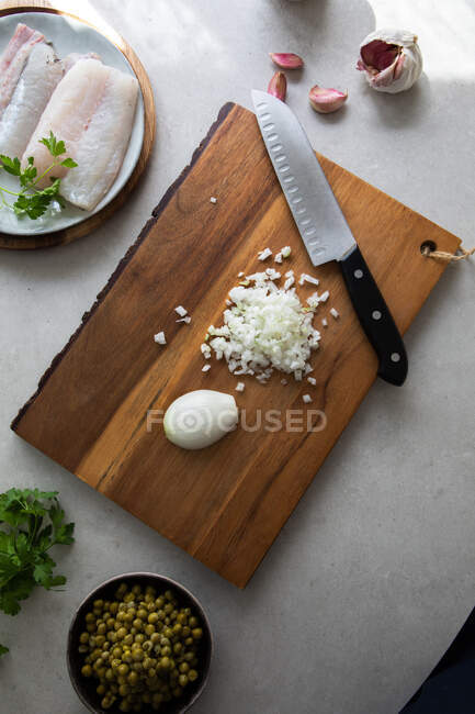 From above wooden board with cut onion and knife placed near peas and hake fillet with herbs during food preparation in kitchen — Stock Photo