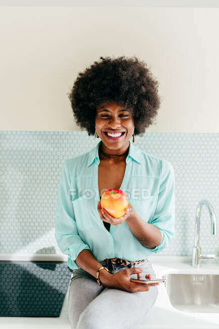 Moderne schöne Afroamerikanerin mit Smartphone in der Hand sitzt auf dem Küchentisch und schaut in die Kamera, lächelt zu Hause und isst Apfel — Stockfoto