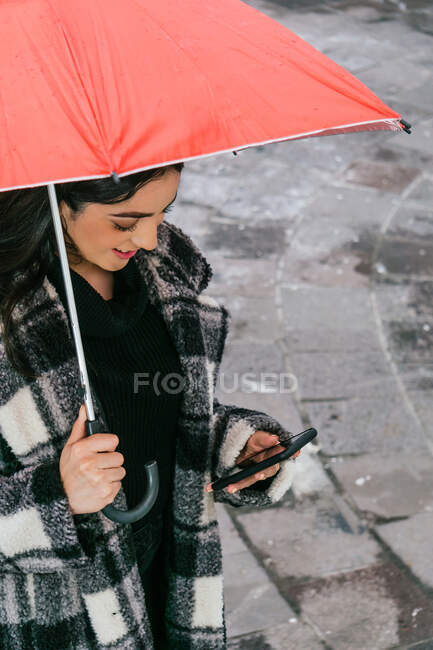 Зверху молода етнічна жінка в картатому пальто переглядає мобільний телефон, стоячи під червоною парасолькою в дощовий день на мокрій вулиці — стокове фото