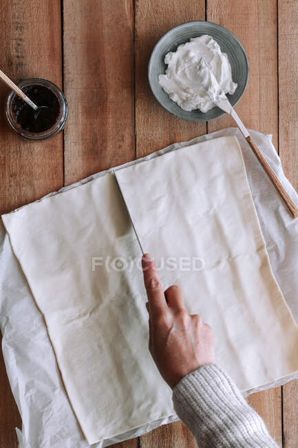 Вид на анонимного человека, разрезающего тонкое тесто на деревянном столе рядом со сливочным сыром и инжирным вареньем во время приготовления выпечки — стоковое фото