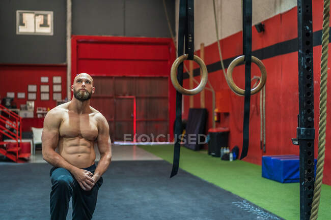 Мускулистый бородатый мужчина смотрит вверх, стоя рядом с оборудованием во время тренировки в современном тренажерном зале — стоковое фото