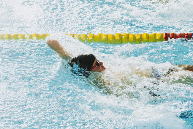 Deportista paralímpico con gafas y gorra sin mano nadando en la piscina entre salpicaduras - foto de stock