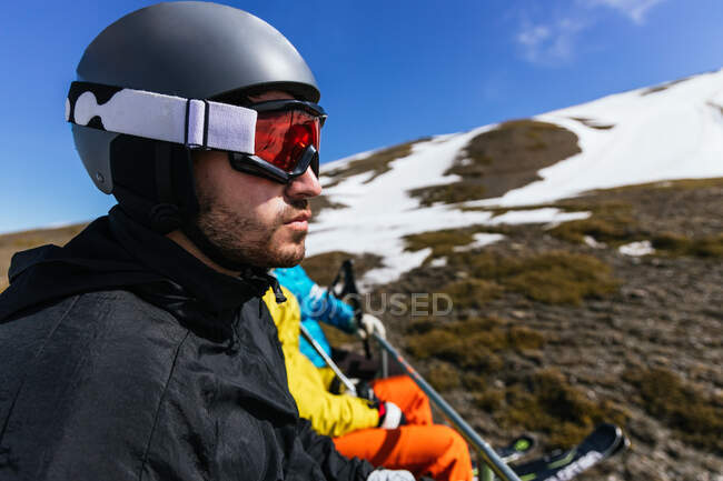 Atleta masculino barbudo en gafas deportivas y casco admirando la montaña de invierno contra socios anónimos en el día soleado en España - foto de stock
