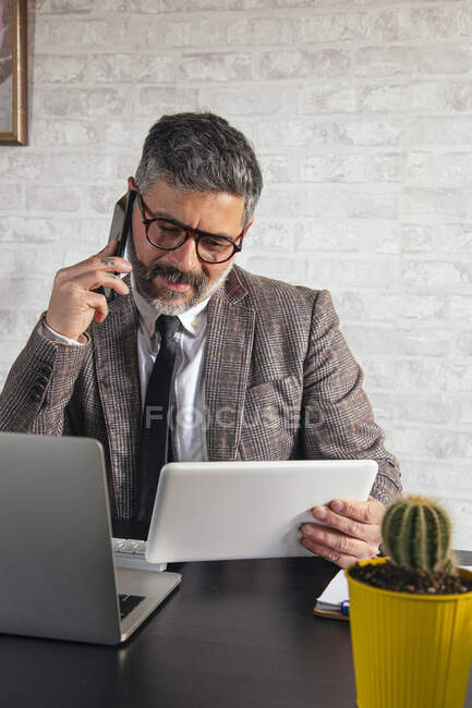 Mature male entrepreneur with tablet talking on cellphone at table with netbook in loft style office — Stock Photo