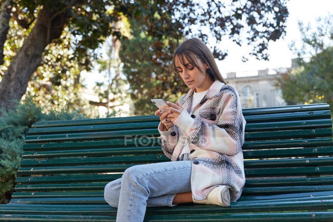 Moderna hembra milenaria en elegante atuendo de primavera sentada en el banco y navegando en el teléfono móvil mientras descansa en la calle urbana mirando hacia otro lado - foto de stock