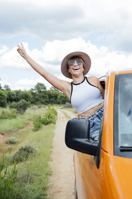 Cheerful female traveler peeping out of van window and showing two fingers gesture while enjoying road trip in summer — Stock Photo