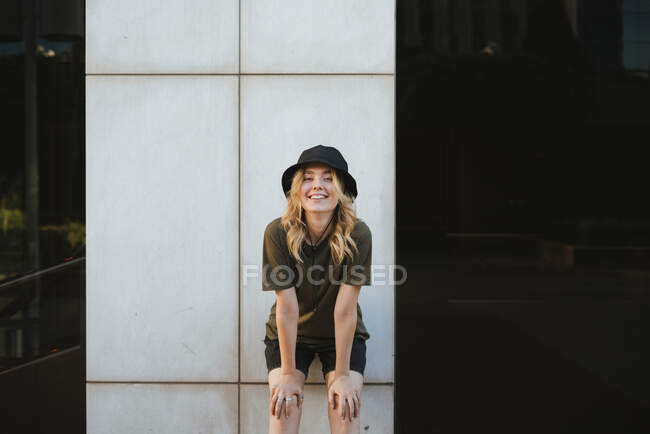Souriante jeune femme en tenue décontractée penchée vers l'avant tout en regardant la caméra en ville en plein jour — Photo de stock