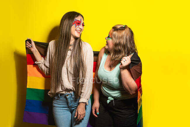 Fidanzate multirazziali con trucco creativo dimostrando bandiera LGBT contro sfondo giallo — Foto stock